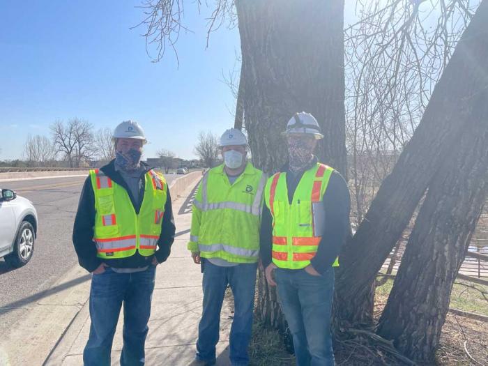 丹佛水安全帽和安全背心的三名男子在路的一边站立在一棵树前面。