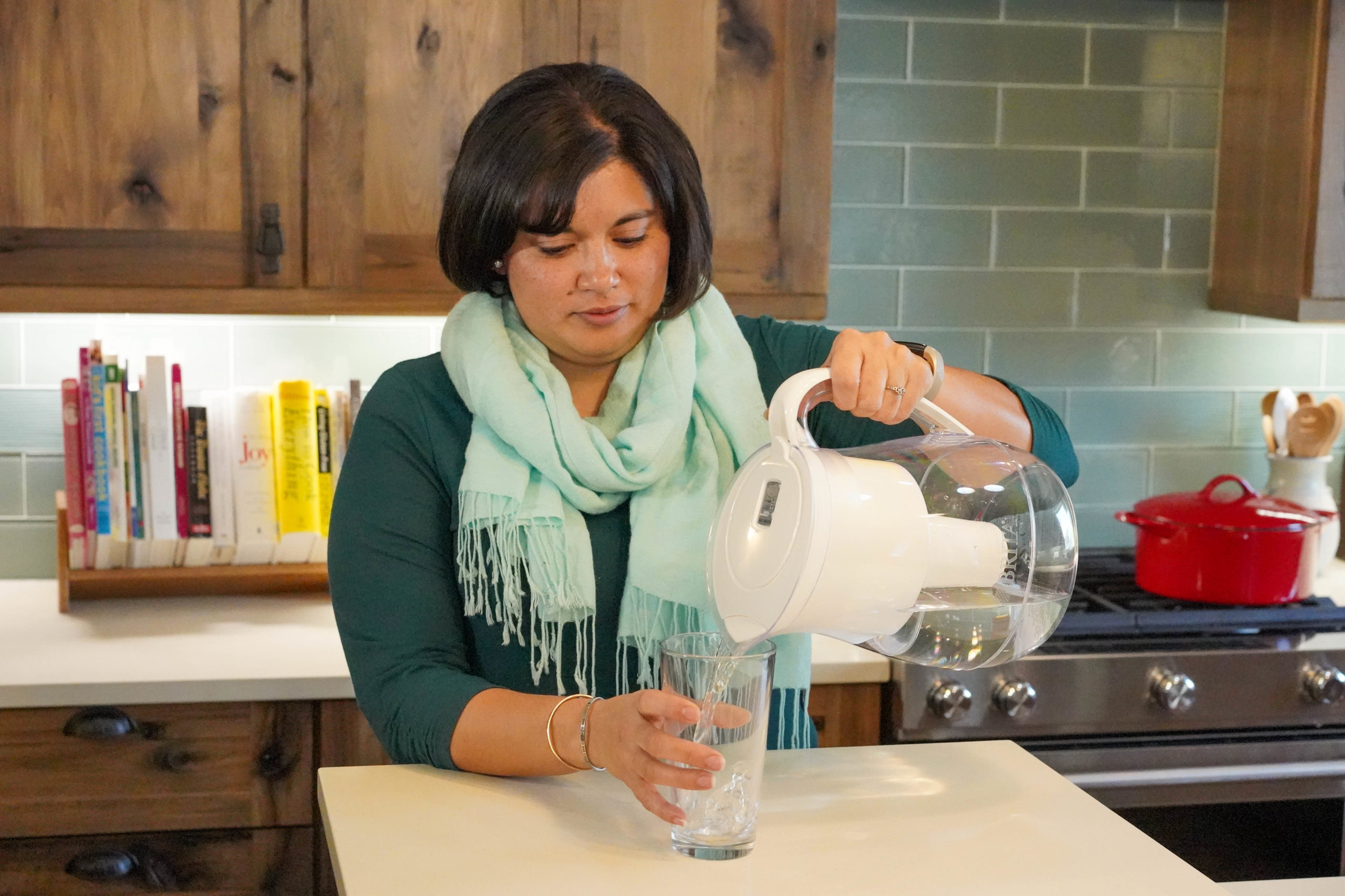一个女人站在一个厨房柜台，从投手和滤水器倒入玻璃杯里喝水。